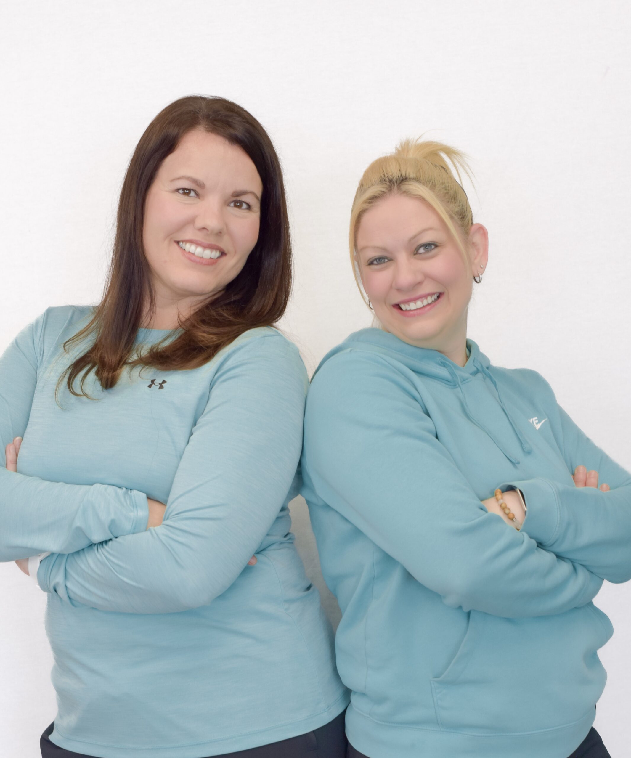 Deux femmes, bras croisés, grand sourir, chandails turquoises, fond blanc